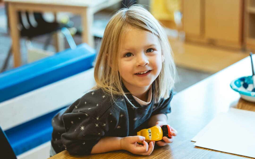 Girl smiling at camera holding toy cars in the classroom at the Early Learning Center